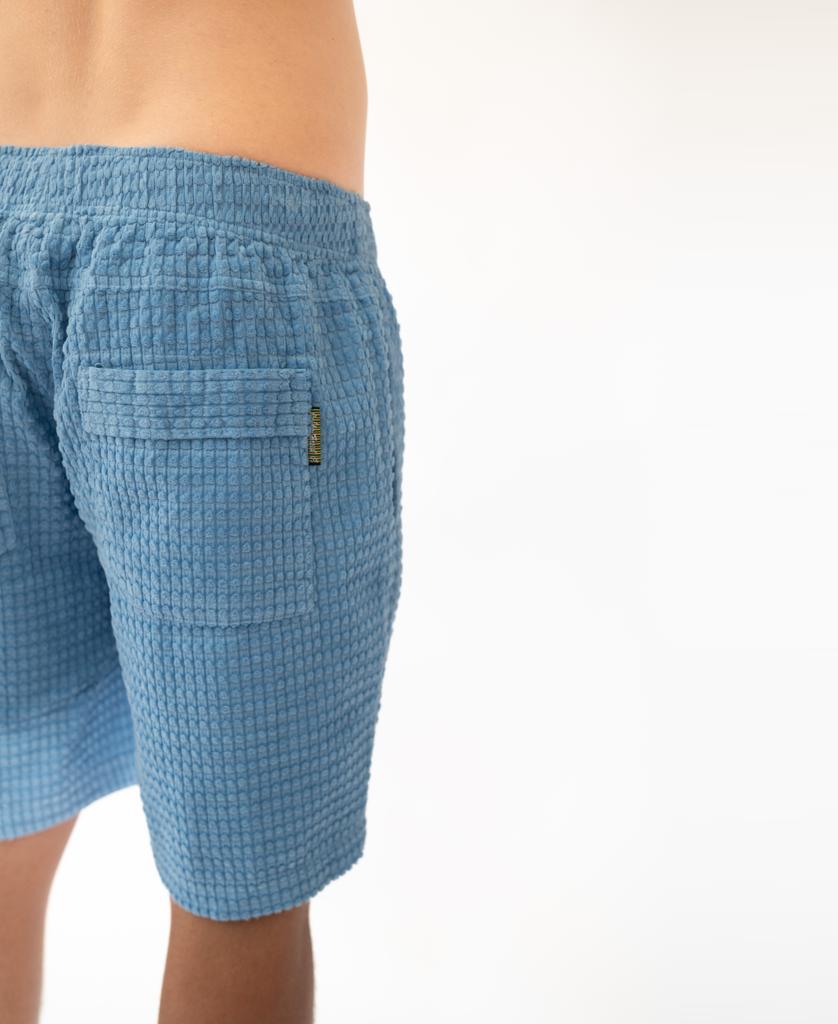 A person wears Golden Breeds El Solito Walkshort in Azure Blue, crafted from soft cotton cord with a visible back pocket, shown waist down against a plain white background.