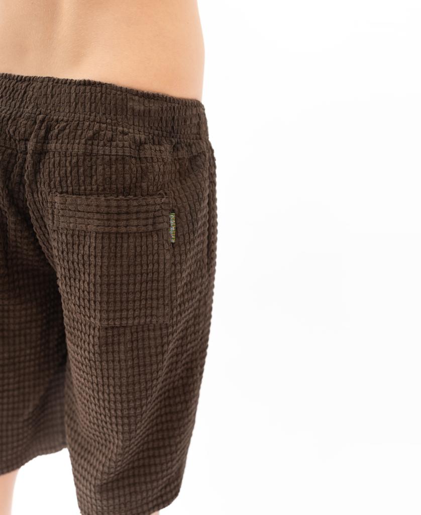 A person wearing Golden Breeds El Solito Walkshort in Wash Brown, made from chunky cotton cord with a textured brown finish and back pocket, stands against a plain white background.