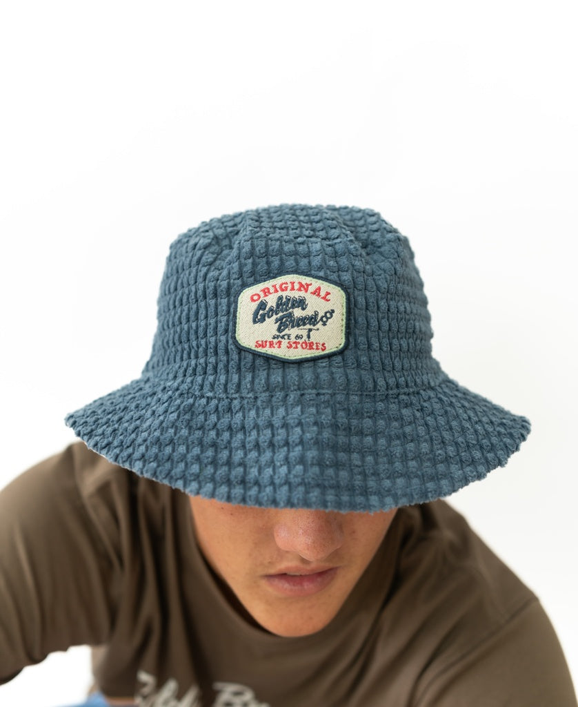 A person wearing the El Noggino Cord Bucket Hat | Denim by Golden Breed, featuring a textured blue fabric and a patch reading Original Clothing Brand & Surf Store. Their face is partially obscured, and they are dressed in a brown shirt.