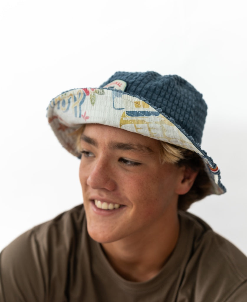 A young person with blond hair smiles and looks to the side against a white background, wearing a brown shirt and the Golden Breed El Noggino Cord Bucket Hat in Denim.