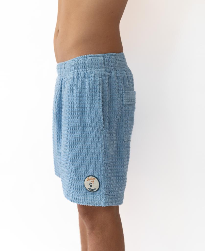 Model wearing Golden Breed El Shorto Walkshort in azure blue, featuring a textured chunky cotton cord, elastic waistband, and a small round patch on the left leg, shown from the side against a white background.