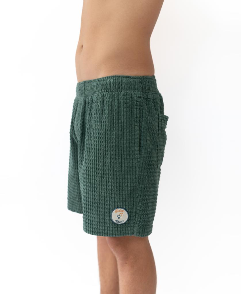 A person in profile wears the Golden Breed El Shorto Walkshort in Ivy Green—elastic waist chunky cotton cord shorts with a textured finish and a round patch near the hem—set against a plain white background.