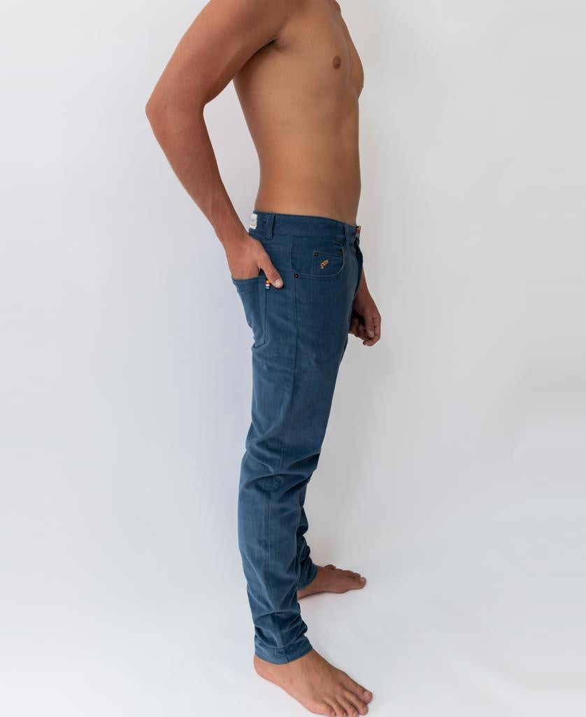 A shirtless person stands barefoot in Felix Stretch Cord Pant | Dusty Blue by Golden Breed, shown in profile with one hand in their pocket against a plain white background.