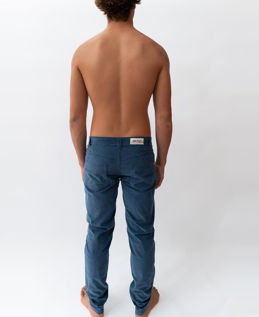 A barefoot person with short hair stands shirtless, facing away from the camera, wearing Golden Breeds Felix Stretch Cord Pant in Dusty Blue against a plain white background.