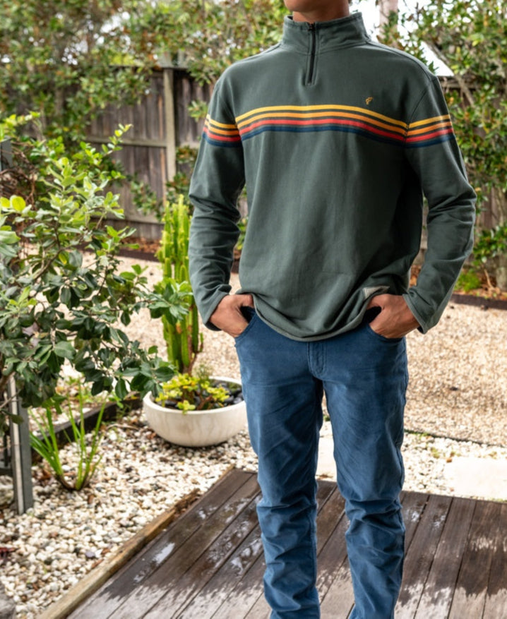 A person stands on a wooden deck, hands in pockets, wearing a green pullover with colorful stripes and Golden Breed’s Felix Stretch Cord Pant in Dusty Blue, surrounded by plants and a gravel garden.
