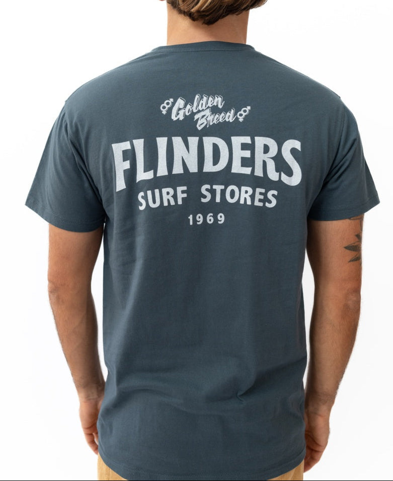 Someone wears the Golden Breed Flinders Easy Rider Tee | Denim, a 100% cotton jersey shirt featuring Golden Breed FLINDERS SURF STORES 1969 in white on the back from the classic Location Tees collection.