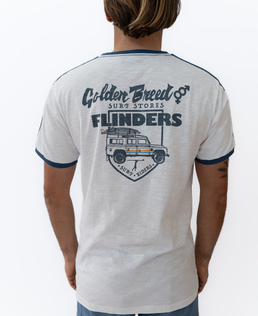 A person wearing the Golden Breed Flinders Landy Tee in Off White—a classic cotton T-shirt with blue trim, FLINDERS text, and a vehicle graphic on the back—captures timeless surf style.