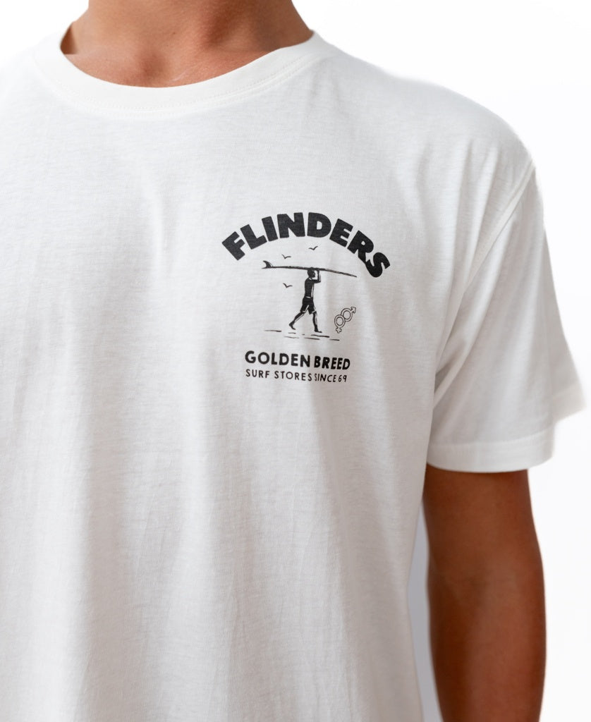 A person wears the Flinders Shanty Tee in Off White by Golden Breed, featuring a FLINDERS GOLDEN BREED SURF STORES SINCE 69 logo with a surfer and birds graphic.