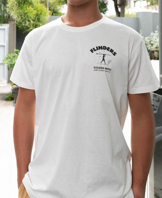 A person wearing the Golden Breed Flinders Shanty Tee in Off White, featuring a small left chest graphic, stands outdoors near a surf store.