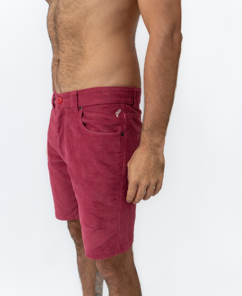 A man wearing Golden Breeds Floyd Cord Walkshort in Scarlet, featuring a structured waist, stands sideways against a plain white background.