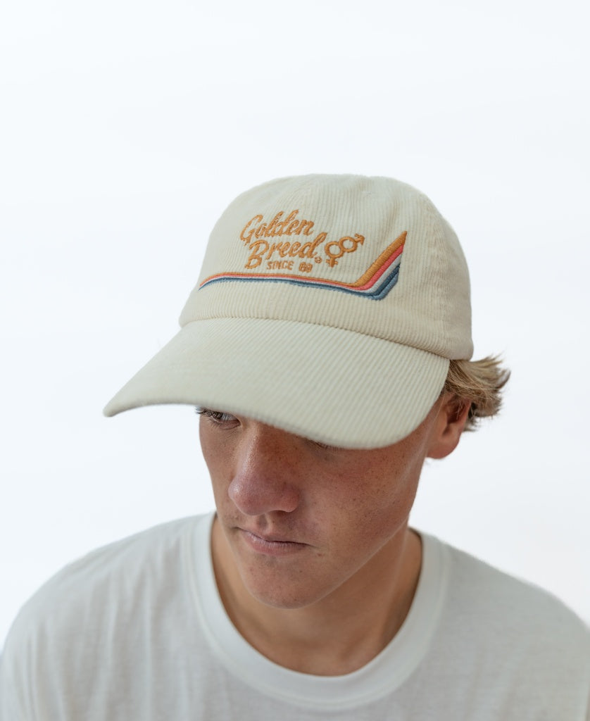 A young person wears a white t-shirt and the Golden Breed Hancock Cord Cap in Off White, featuring Since 69 and a rainbow design embroidered on the front, looking downward.
