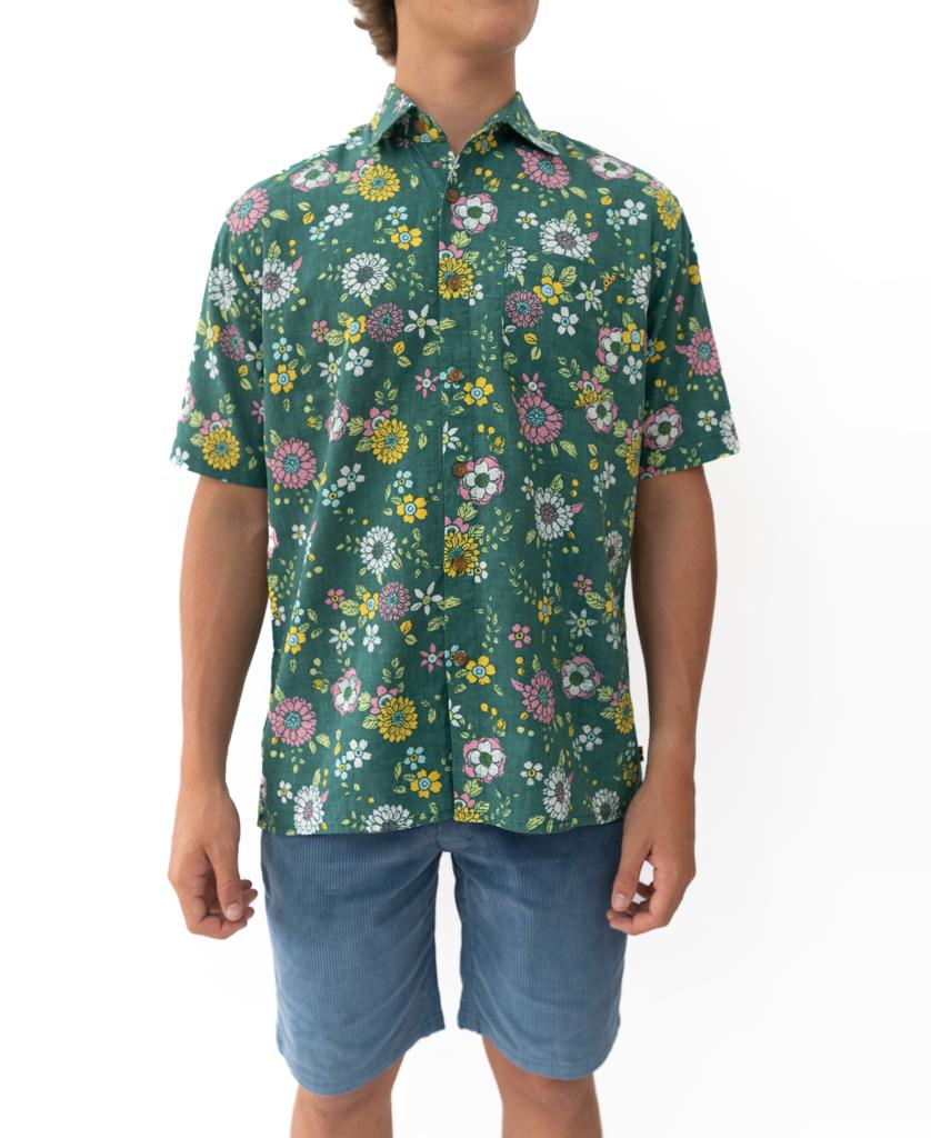 A person wears the Golden Breed Happy Floral | Ivy Green short-sleeve Hawaiian shirt with a colorful floral pattern and light blue relaxed-fit shorts, standing against a white background.