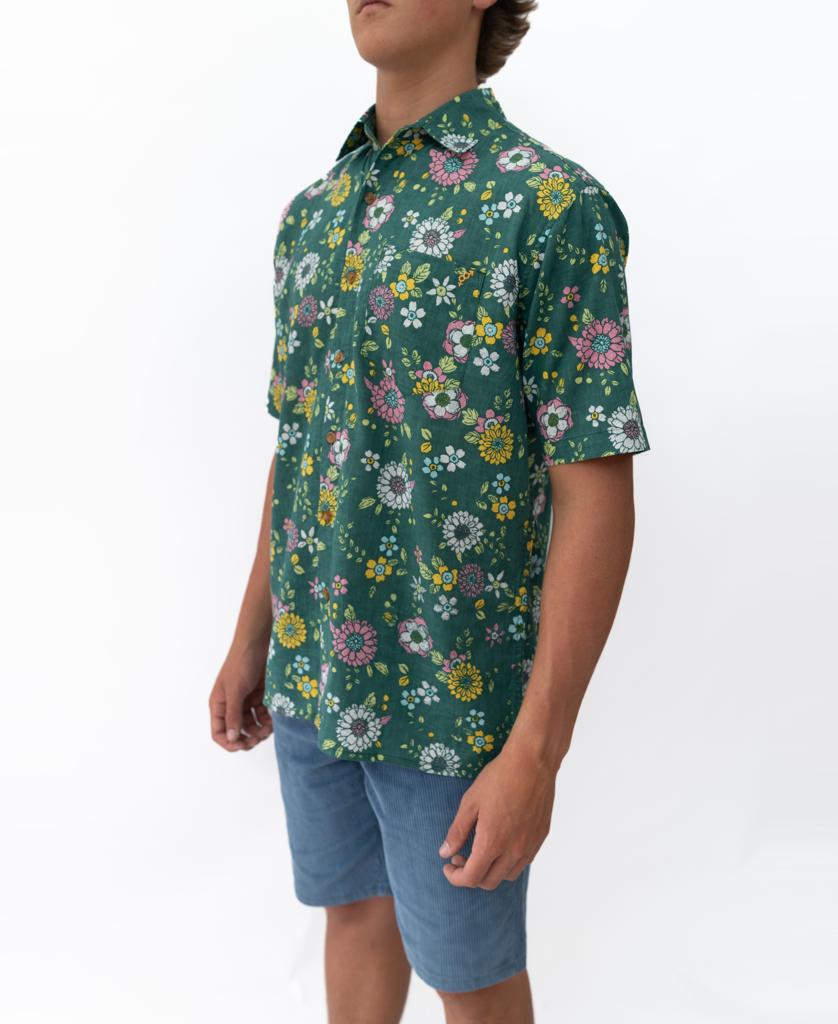 A person wearing Golden Breeds Happy Floral | Ivy Green short-sleeve shirt and blue shorts stands against a plain white background.
