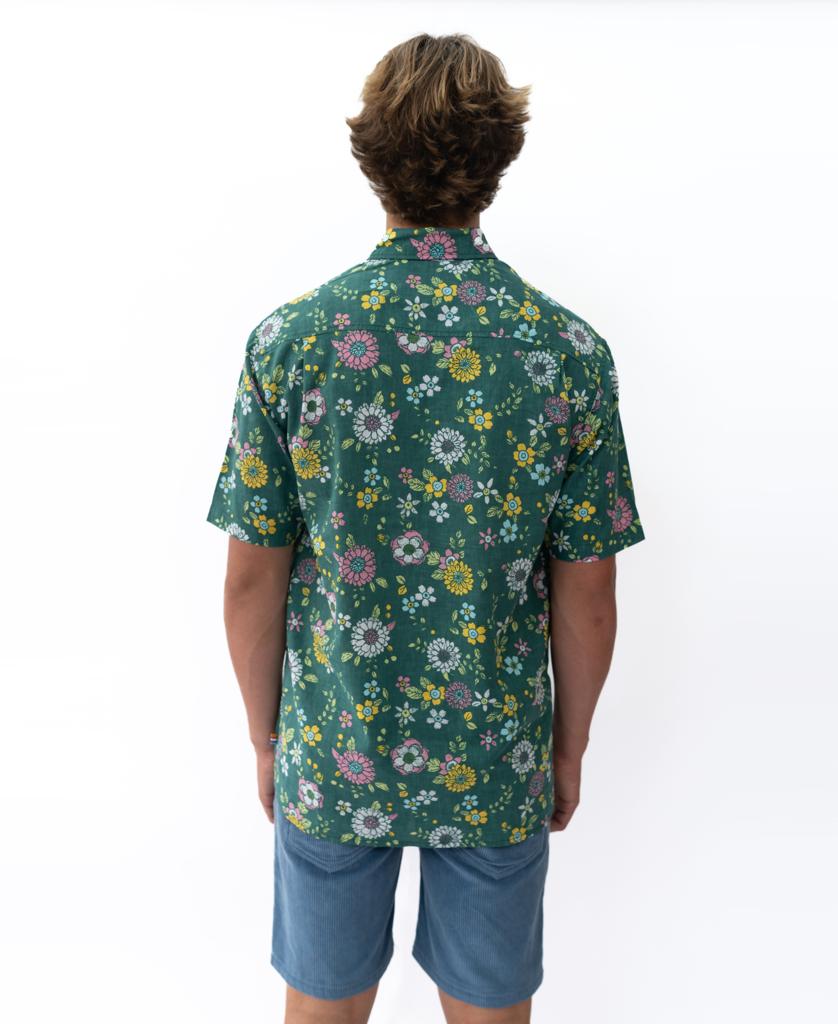 A person stands facing away, wearing the Golden Breed Happy Floral | Ivy Green shirt with a relaxed fit and blue shorts, set against a plain white background.