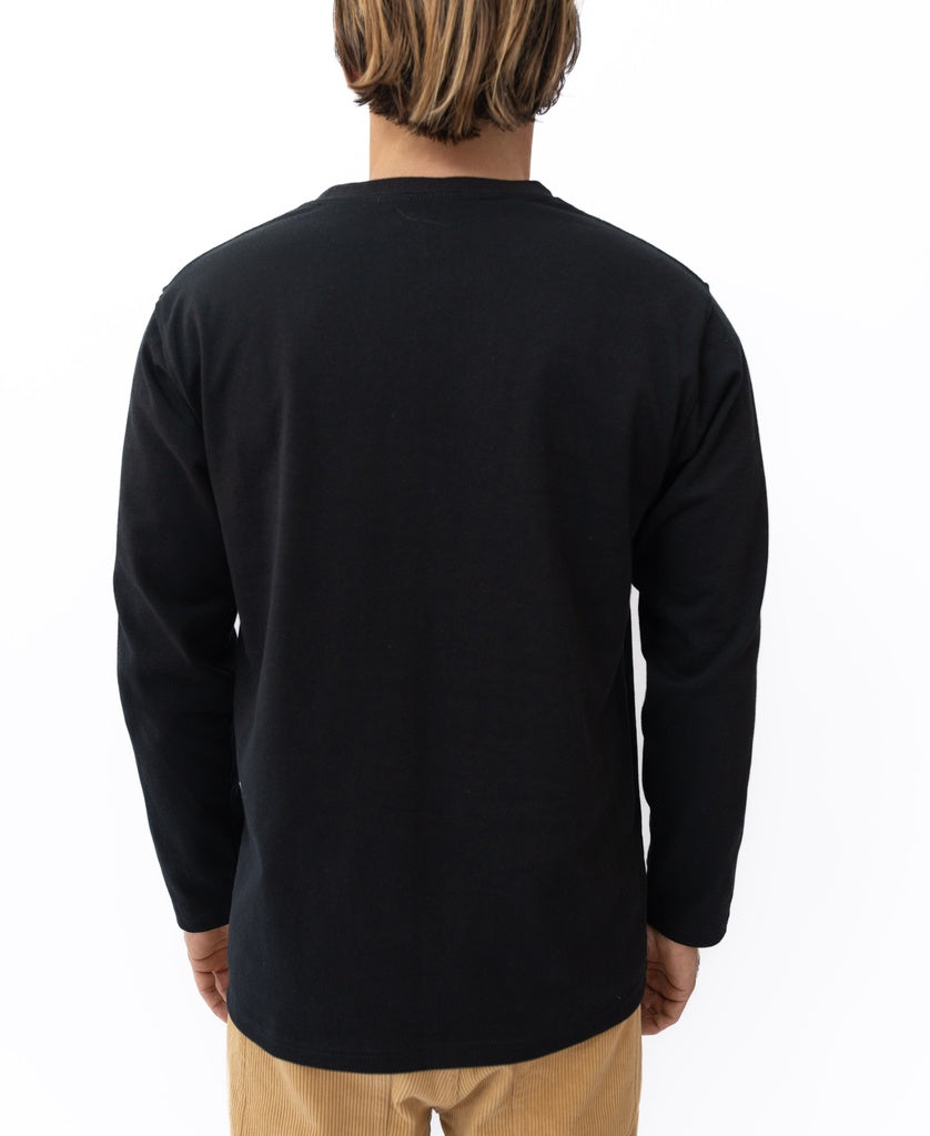 A person faces away from the camera in the Golden Breed Heavy Island Long Sleeve Tee | Black, 100% heavy cotton jersey, paired with light brown pants for a laid-back Cali surf vibe against a plain white background.