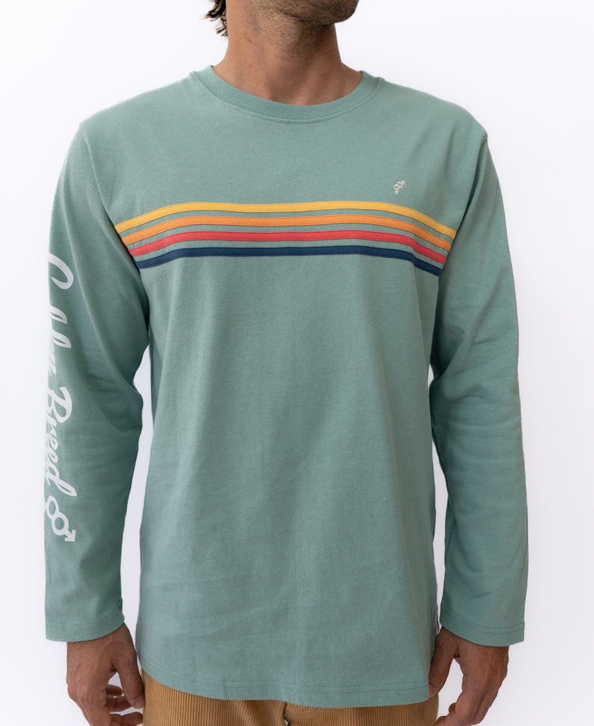 Wearing the Golden Breed Heavy Island Long Sleeve Tee in Sage, with colorful stripes and white sleeve script, a person embodies a laid-back beach surf vibe against a plain white background.