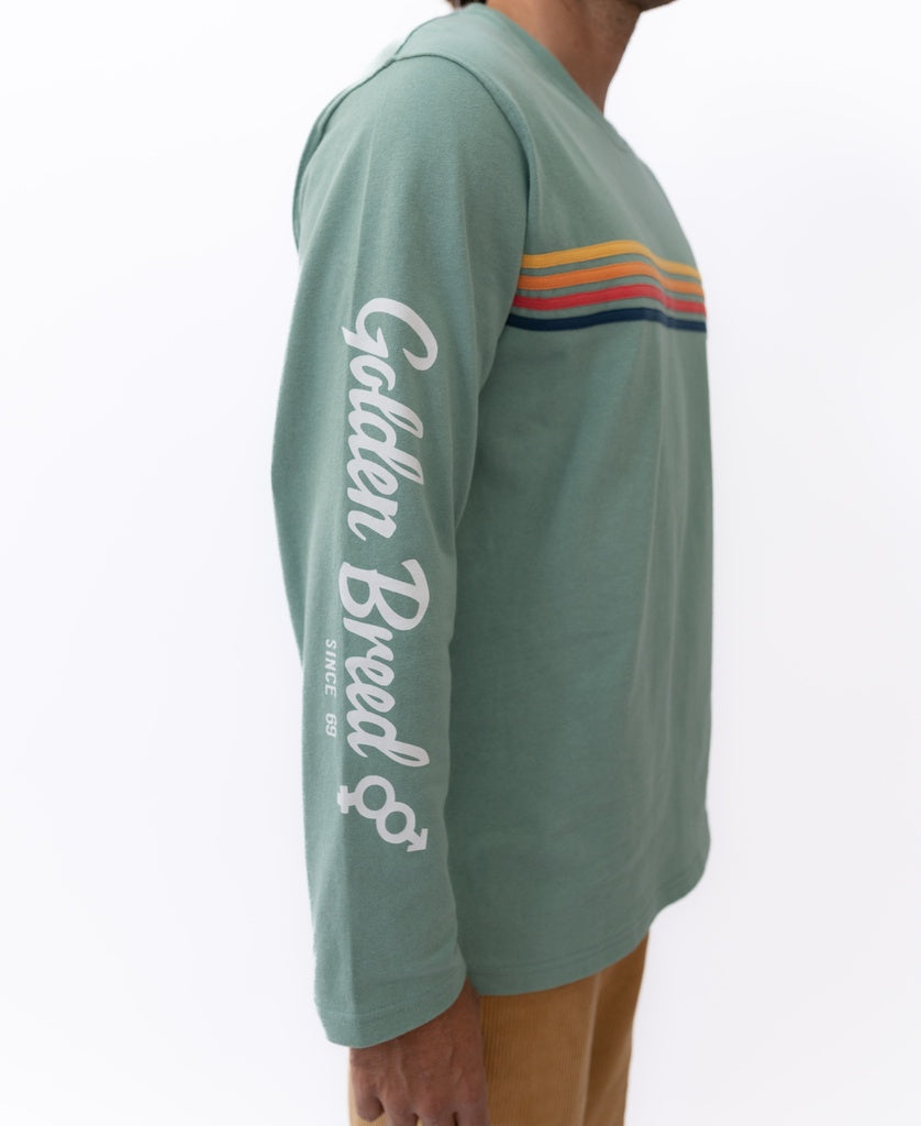 A person wears the Golden Breed Heavy Island Long Sleeve Tee in Sage, featuring colorful stripes and branding on the sleeve, paired with tan pants for a relaxed surf vibe. The plain white background enhances the easy 60’s California style.
