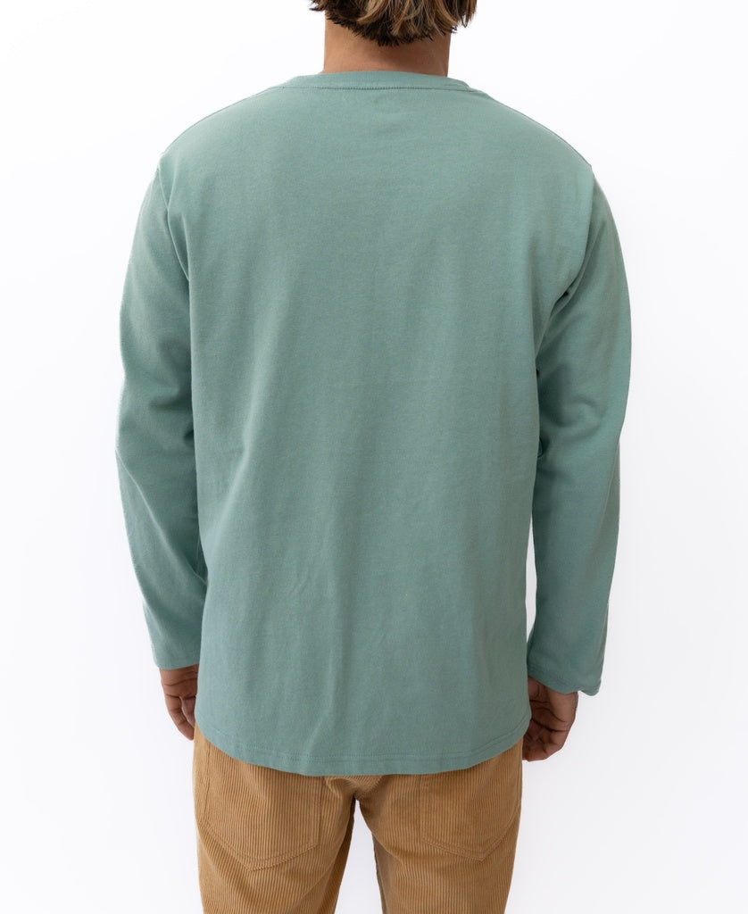 A person stands with their back to the camera, wearing Golden Breeds Heavy Island Long Sleeve Tee in Sage and tan corduroy pants, capturing a laid-back beach surf vibe against a plain white background.