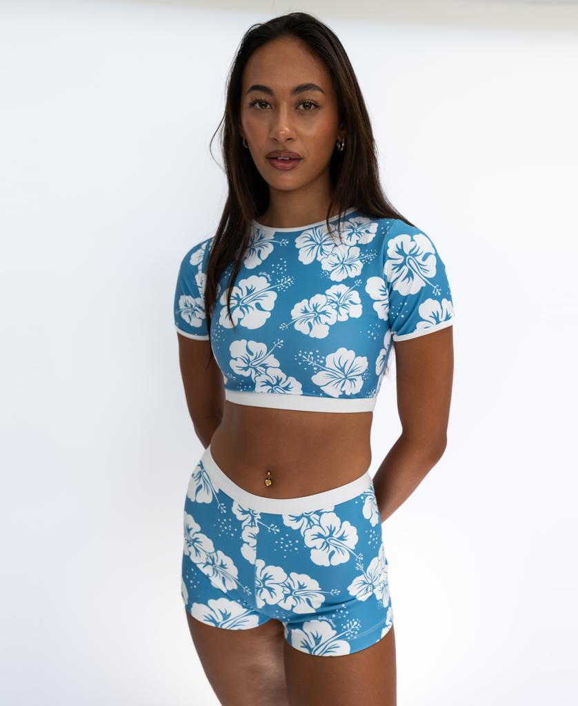 A woman stands against a white background wearing the Golden Breed Indie Crop Tee in Hibiscus Azure / Off White, featuring a blue and white floral print made from eco-friendly recycled fabric.