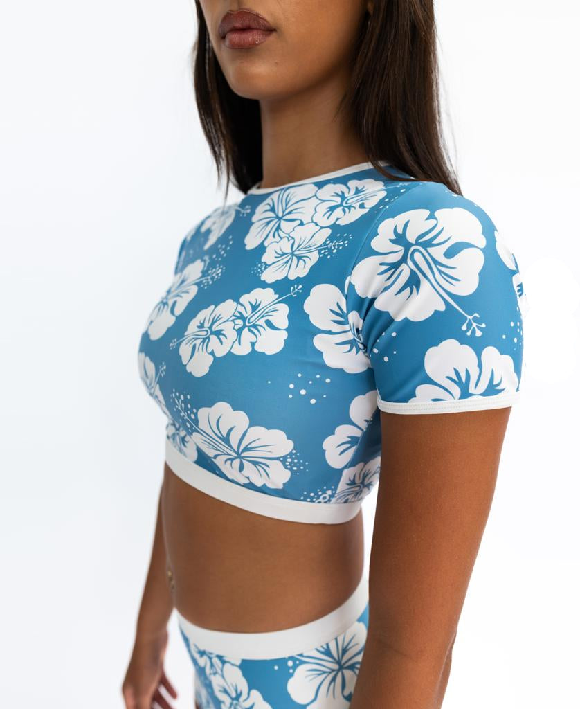 A woman wears the Golden Breed Indie Crop Tee in Hibiscus Azure / Off White, featuring eco-friendly recycled fabric and white hibiscus prints, paired with matching high-waisted bottoms against a plain white background.