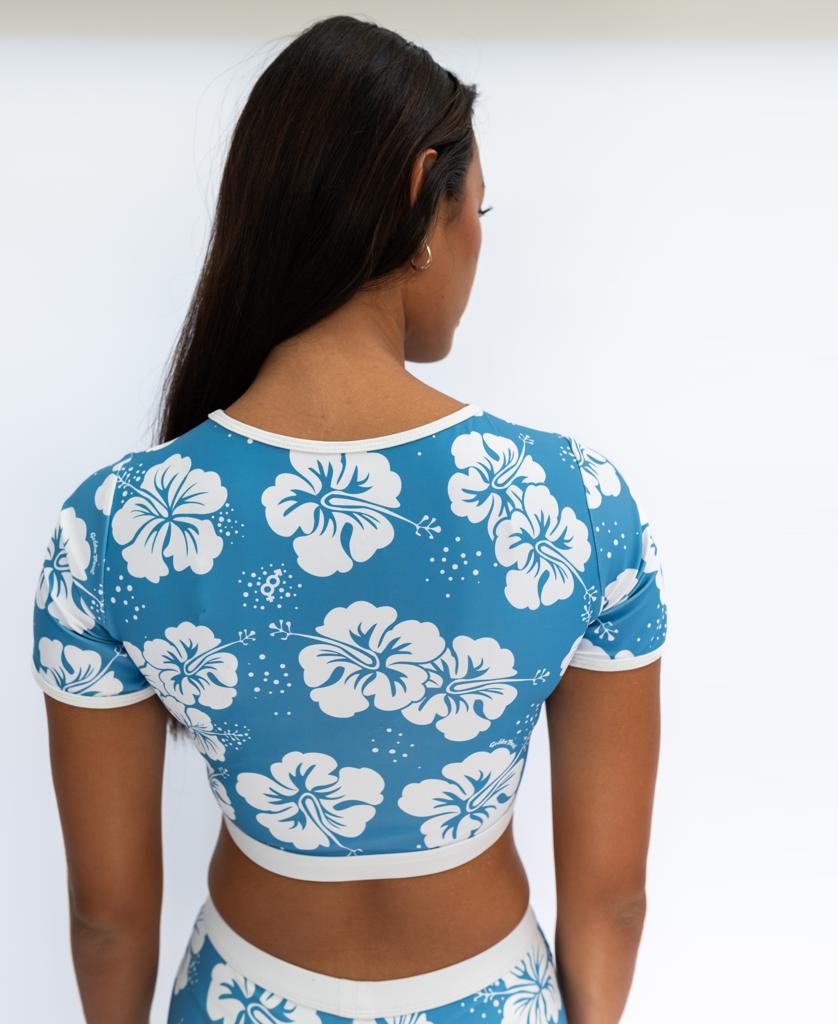 A person with long dark hair faces away, wearing the Golden Breed Indie Crop Tee in Hibiscus Azure/Off White, featuring a white hibiscus pattern and matching bottoms made from recycled fabric, set against a plain white background.