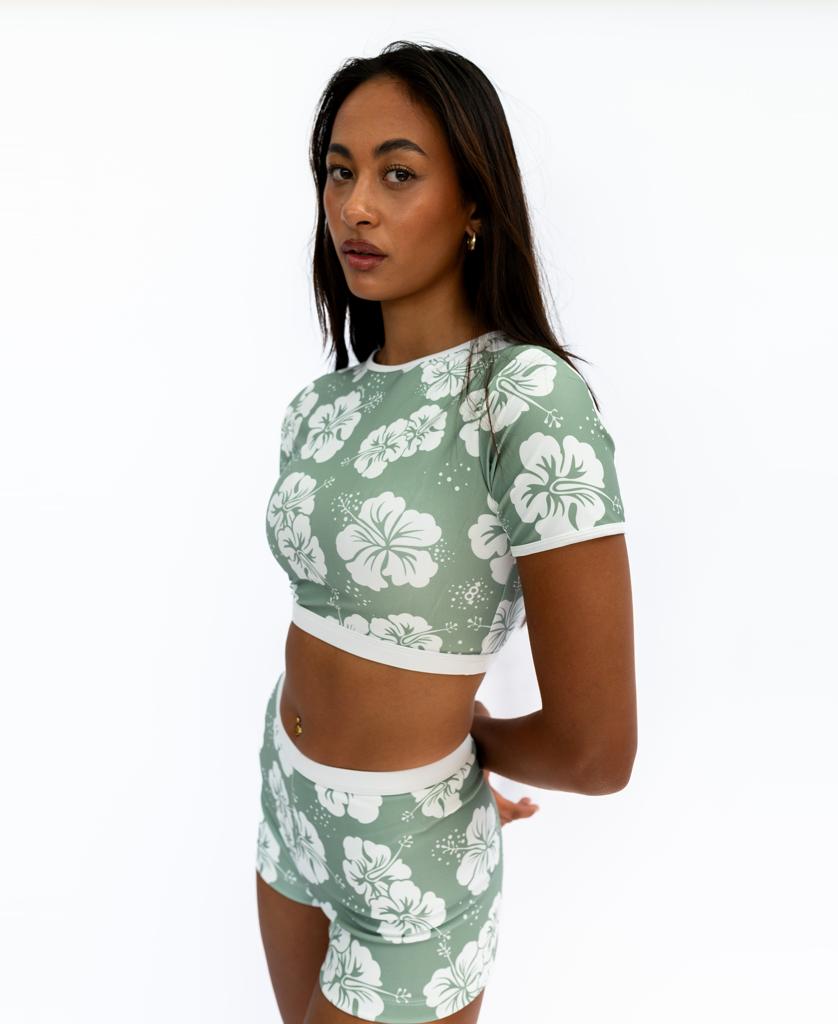 A woman stands against a white background, wearing the Golden Breed Indie Crop Tee in Hibiscus Moss/Off White, an eco-friendly top made from recycled fabric, looking at the camera with her arms behind her back.