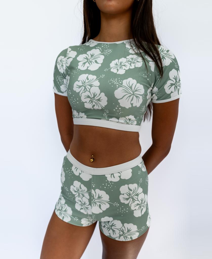 A person wears the Golden Breed Indie Crop Tee in Hibiscus Moss / Off White, featuring a white hibiscus print on green recycled fabric, paired with matching shorts, standing against a plain white background.