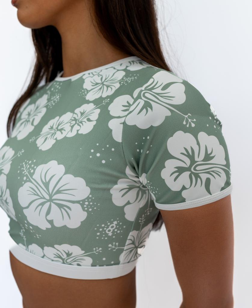 Someone wears the Golden Breed Indie Crop Tee in Hibiscus Moss / Off White, featuring a white hibiscus flower pattern on eco-friendly, recycled fabric, standing against a plain white background.