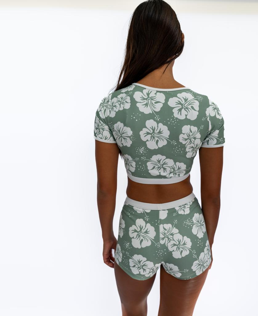 A woman faces away, wearing the Golden Breed Indie Crop Tee in Hibiscus Moss/Off White with white hibiscus prints, made from recycled fabric, paired with matching shorts, set against a plain white background.
