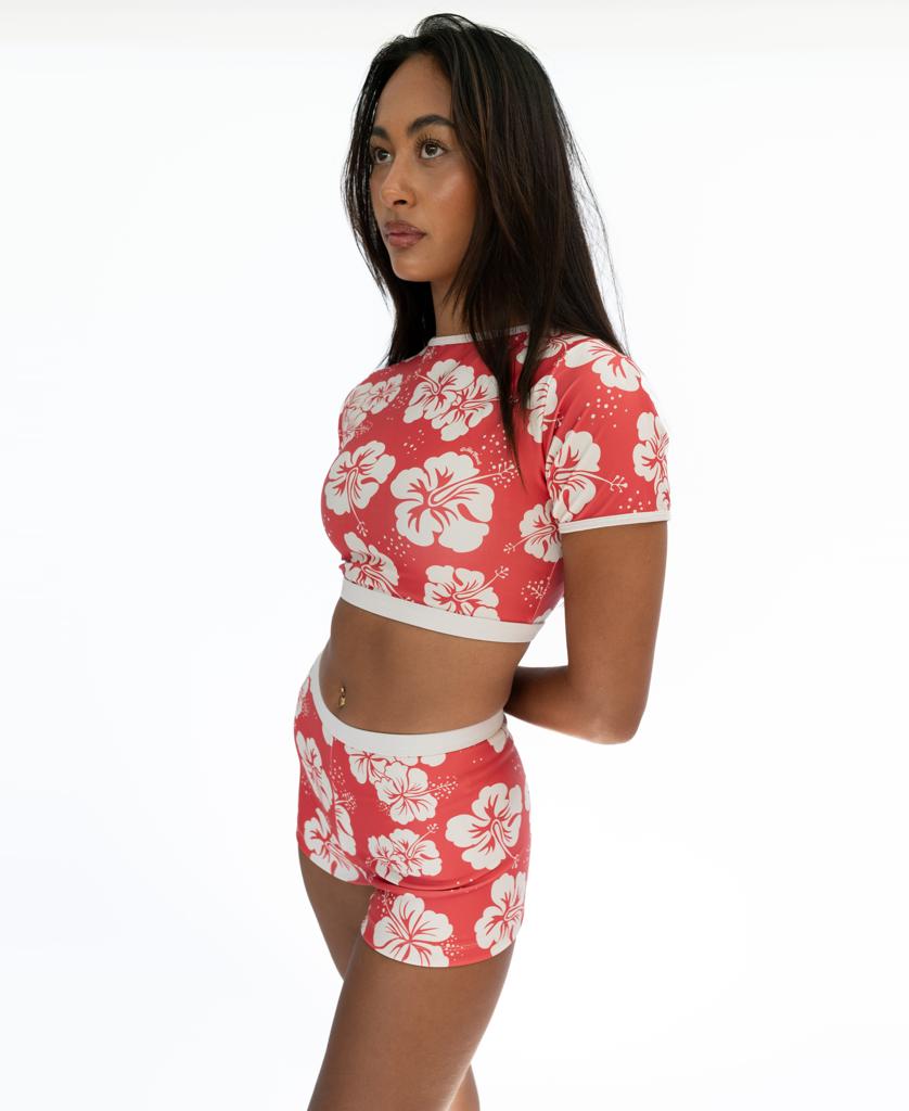 A woman in profile wears the Golden Breed Indie Crop Tee in Hibiscus Wash Red/Beige, featuring white hibiscus prints and made from eco-friendly recycled fabric, paired with matching shorts against a plain white background.