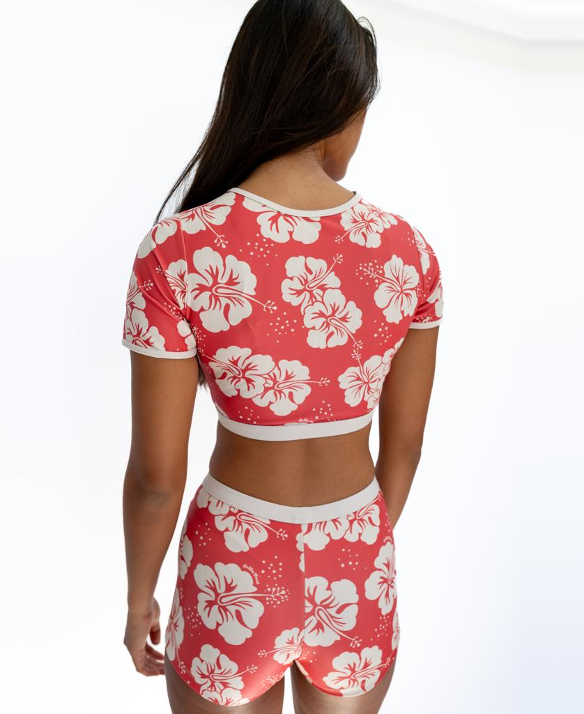 A woman faces away, wearing the Golden Breed Indie Crop Tee in Hibiscus Wash Red / Beige, made from recycled fabric, against a plain white background.