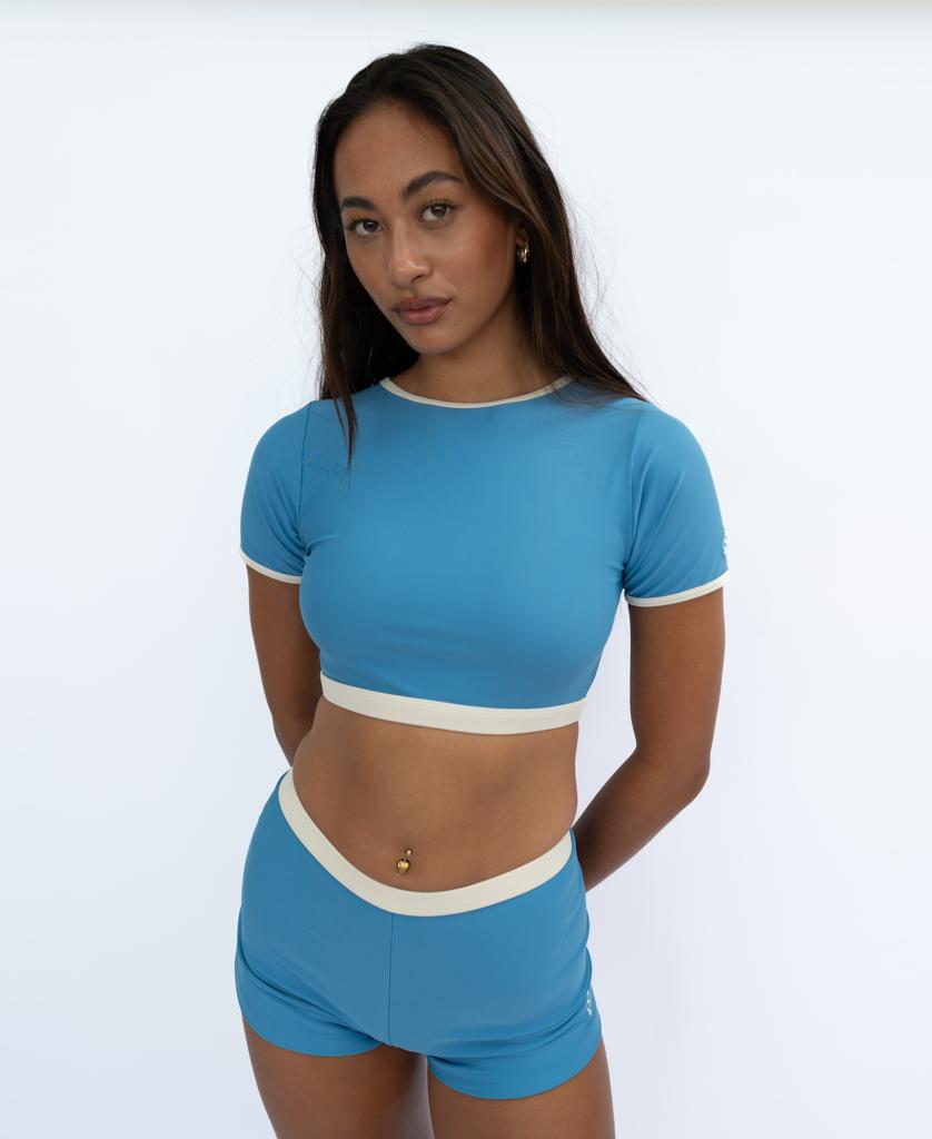 A woman stands against a plain white background in the Golden Breed Indie Crop Tee Retro (Azure/Off White)—an effortless tribute to iconic surf style.