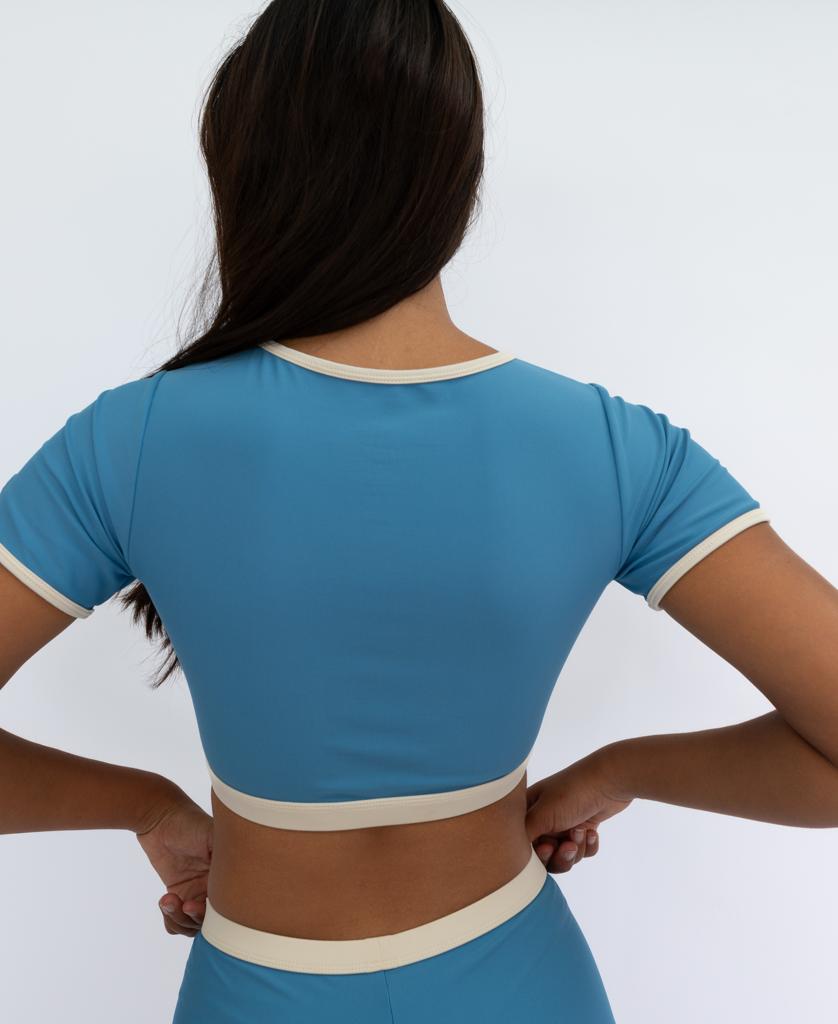 A person stands with their back to the camera, wearing the Golden Breed Indie Crop Tee Retro in Azure and Off White, capturing a vintage-inspired beach vibe.