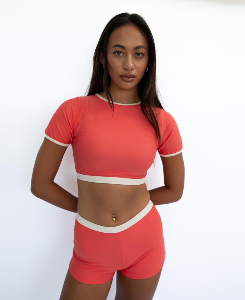 A woman stands against a white background wearing the Golden Breed Indie Crop Tee Retro in Surf Red/Off White, hands behind her back and a neutral expression, evoking retro surf vibes.