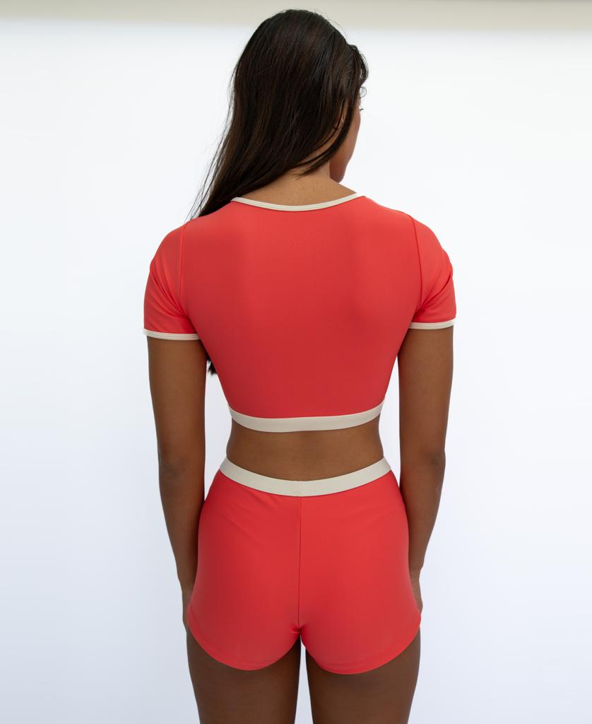 A woman stands with her back to the camera wearing the Golden Breed Indie Crop Tee Retro in Surf Red / Off White, paired with matching high-waisted shorts, both from the brands surf swim range, against a plain white background.