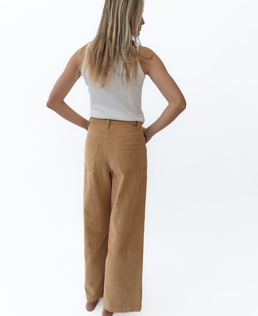 A woman with long blonde hair wears a white sleeveless top and Golden Breed Jenny Cord Pants in camel, photographed from the back against a plain white background.