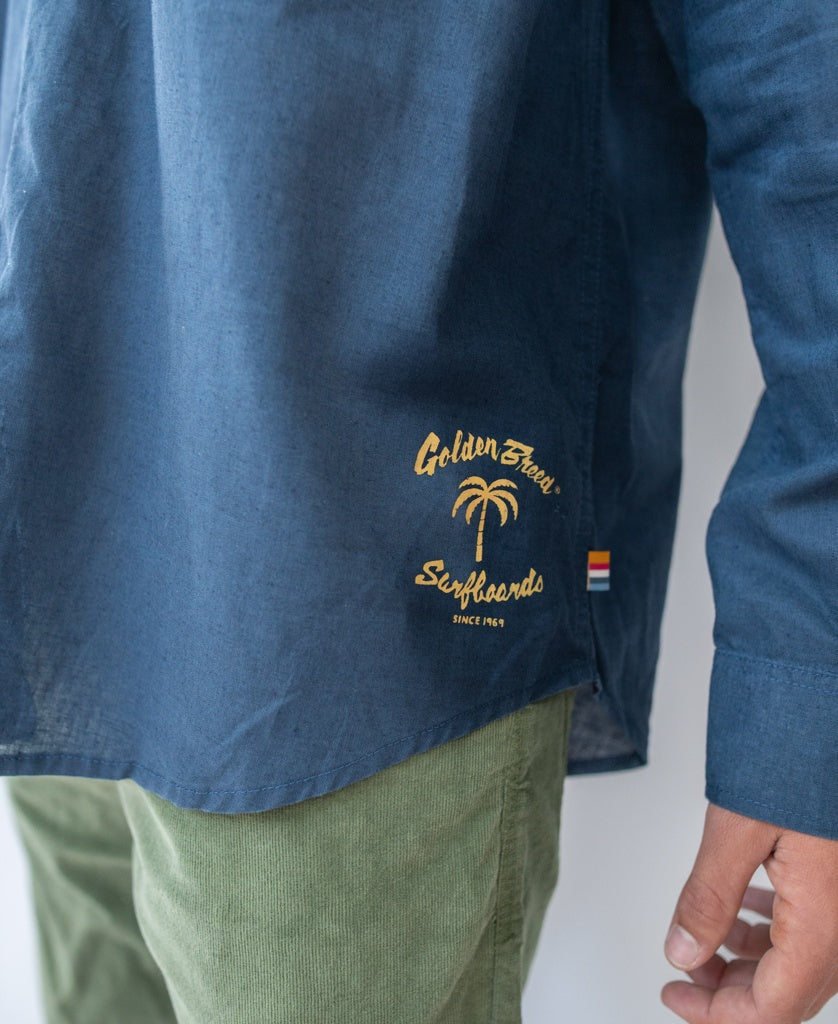 A person wears the Golden Breed Linen LS Shirt in Vintage Navy, featuring a Golden Sands Surfboards and palm tree graphic, paired with green pants. A small rainbow tag accents the shirt.