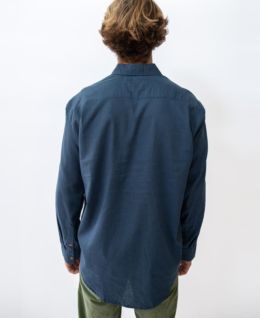 A person with light brown hair faces away from the camera, wearing Golden Breed’s Linen LS Shirt in Vintage Navy and green pants, set against a plain white background.