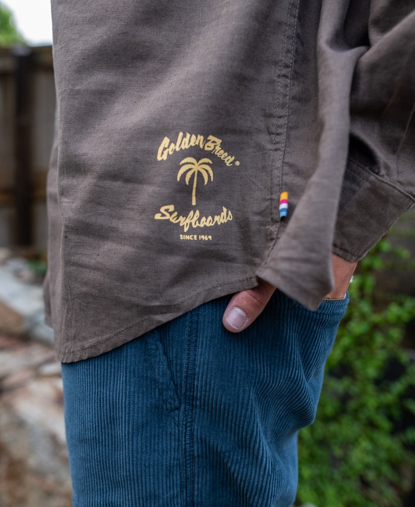 A person wearing blue corduroy pants and a brown Linen LS Shirt by Golden Breed, featuring Golden Bear Surfboards Since 1969 and a palm tree logo.
