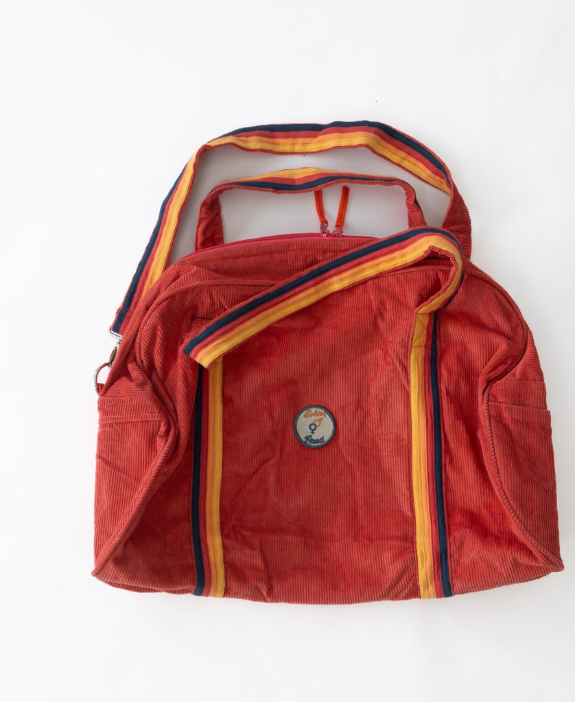 The Golden Breed Long Island Cord Roll Bag in Wash Red is crafted from cotton corduroy, features yellow and blue striped handles and trim, and a front round patch with an illustration and Golden Breed text.