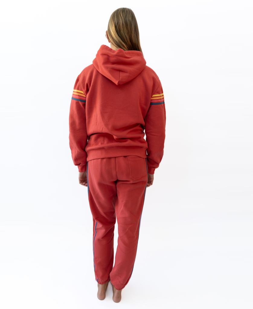 A person stands barefoot facing away, wearing the Golden Breed Long Island Hoodie in Wash Red with striped sleeves and matching sweatpants, all crafted from 100% Cotton Fleece, set against a white background.