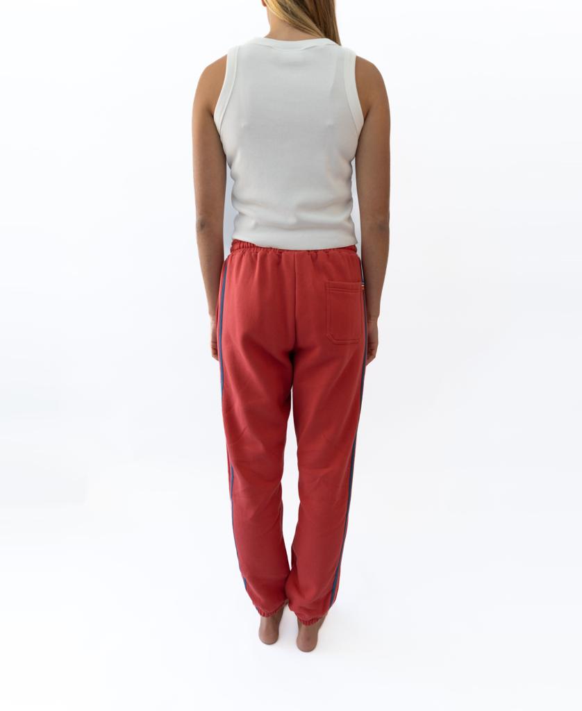 A person stands barefoot, facing away, wearing a white sleeveless top and Golden Breeds Long Island Crunchie T/P trackpant in Wash Red with an elastic waistband and back pocket, set against a white background.
