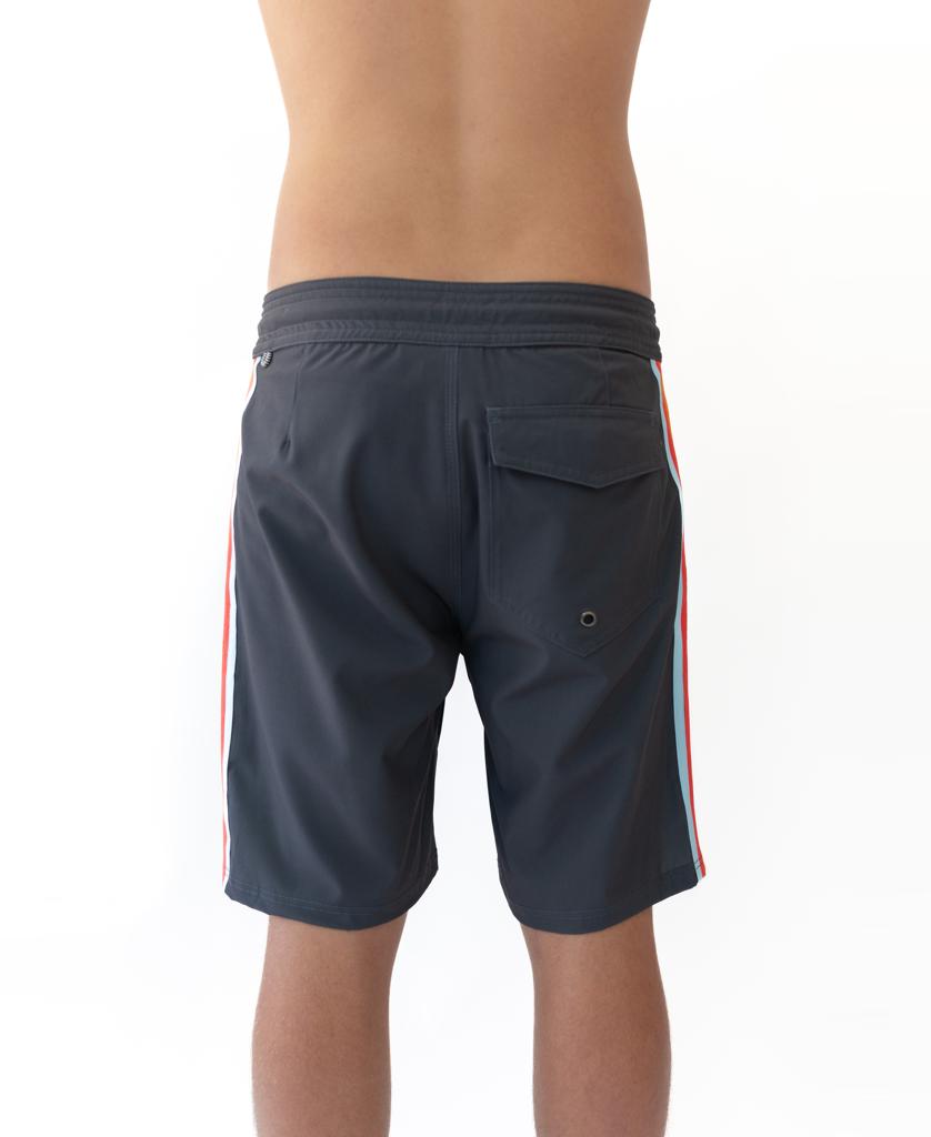 A person stands with their back to the camera in Golden Breeds Long Island Boardie | Wash Black shorts with side stripes and a back pocket, channeling a 60’s California surf vibe against a plain white background.