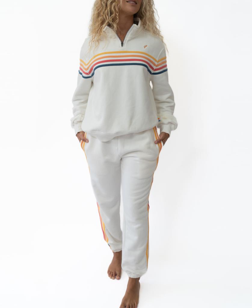 A person stands barefoot against a white background, wearing the Golden Breed Long Island 1/4 Zip in off white with matching joggers, both featuring multicolored stripes and a Retro Golden Breed Logo.