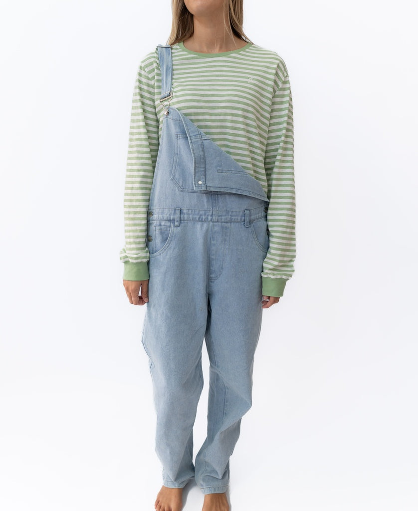 A person in Golden Breeds Maci Overall in light blue, with adjustable straps (one unfastened), and a green and white striped long-sleeve shirt stands barefoot against a white background.