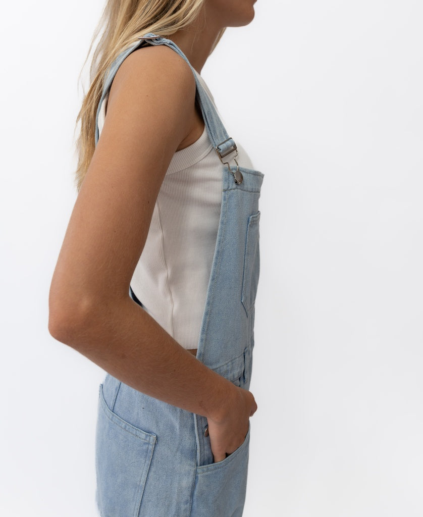 A person with long blonde hair, seen from the side, wears the Golden Breed Maci Overall in Light Blue over a white ribbed tank top, with one hand in the pocket, posed against a plain white background.
