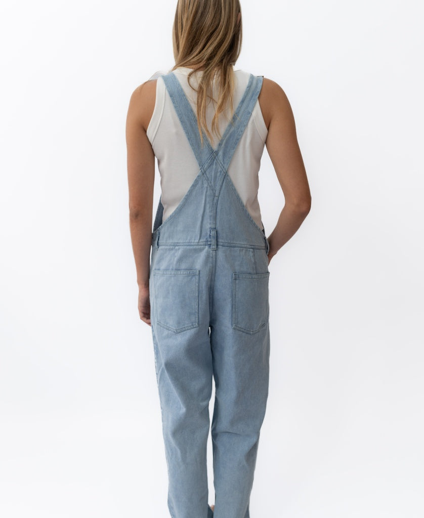 A person stands facing away from the camera, wearing Golden Breeds Maci Overall in light blue with adjustable straps and a white sleeveless top, set against a plain white background.