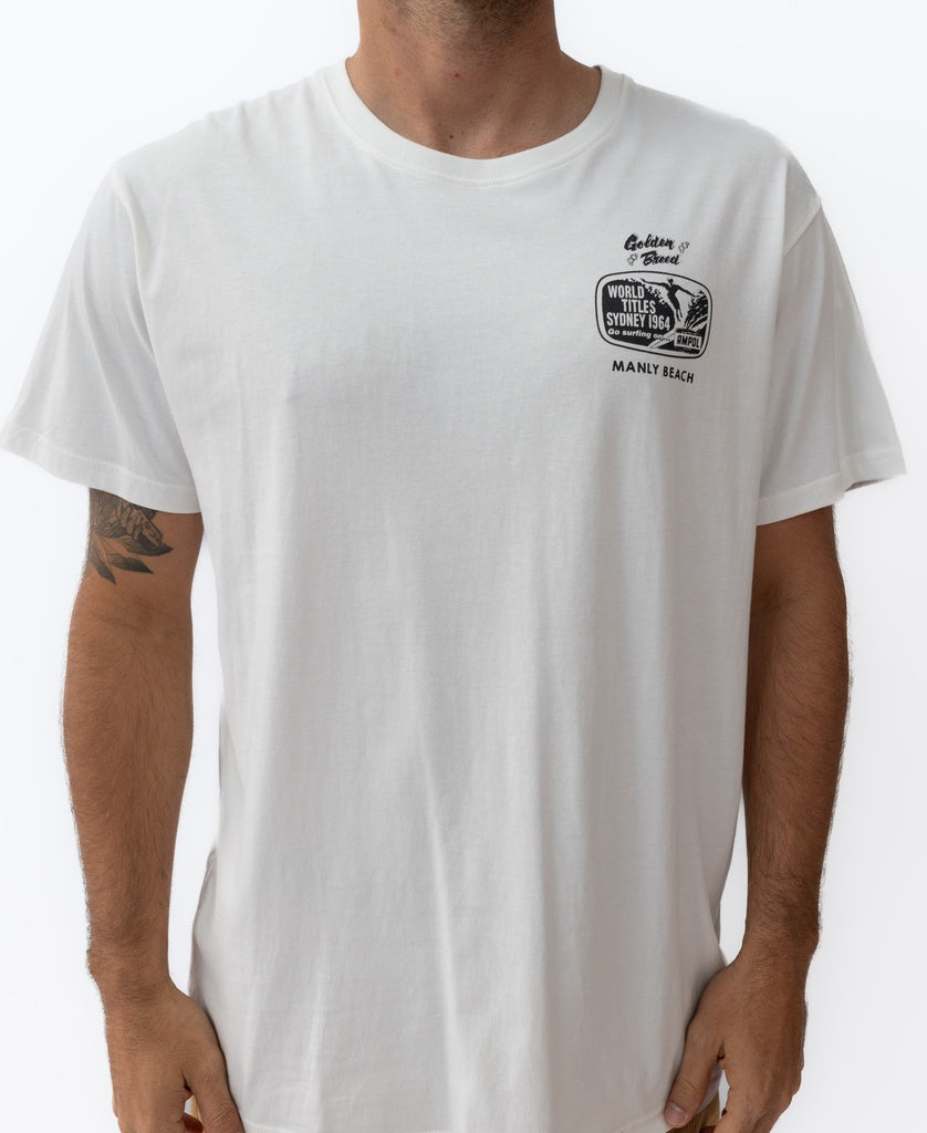 A person wearing the Golden Breed Manly Ampol Tee in off white, featuring a small black graphic and Wylies Store, Manly Beach text on the upper left chest.