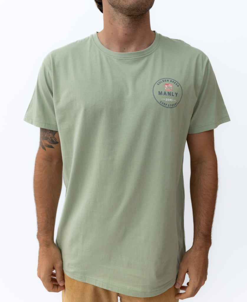 A man wearing the Golden Breed Manly Kombi Tee in Sage, made from 100% cotton and featuring a small printed logo on the left chest, stands against a plain white background.