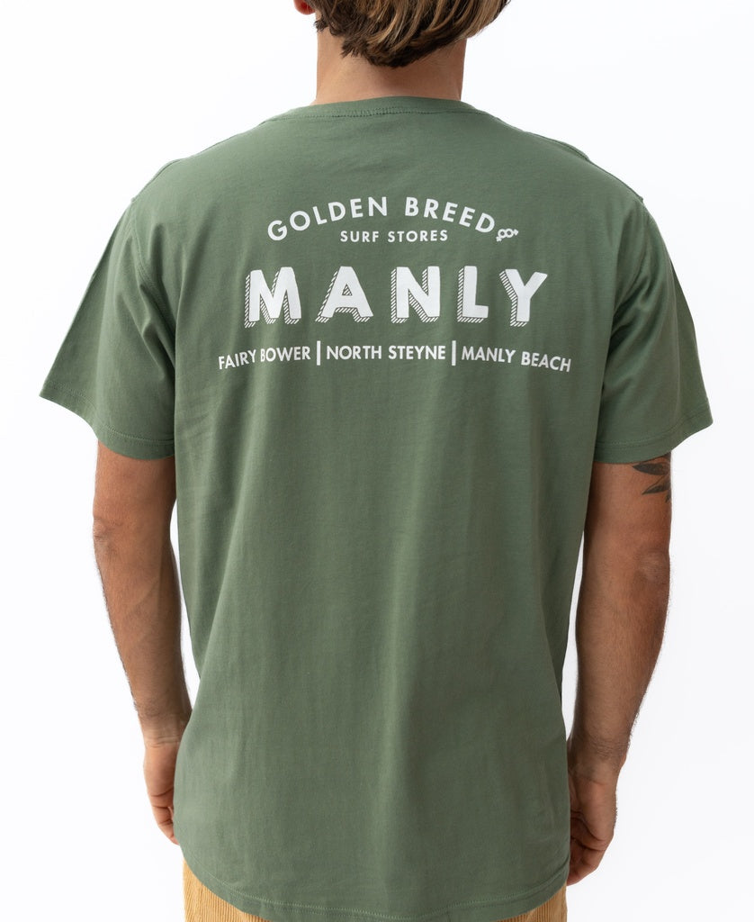 A person wearing a khaki 100% cotton Manly Old School Tee by Golden Breed, featuring Surf Stores MANLY Fairy Bower North Steyne Manly Beach in white text on the back.